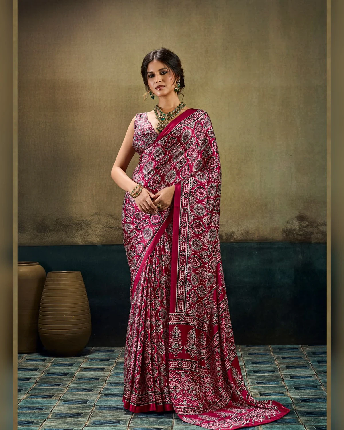 Woman wearing a crimson all-over Ajrakh print modal satin saree, standing by clay pots on a blue tiled floor