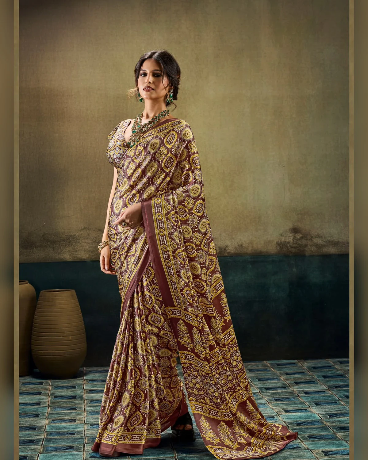Woman wearing a brown all-over Ajrakh print modal satin saree, standing by clay pots on a blue tiled floor