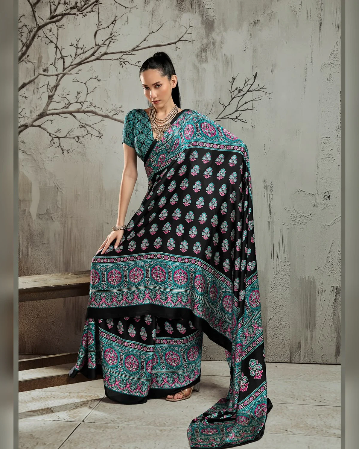 Woman wearing a black and teal Ajrakh buti modal satin saree with a wide teal border, against a grey wall with bare branches
