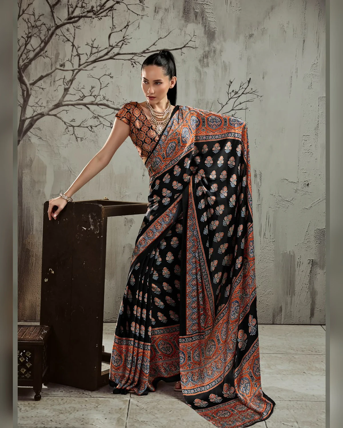 Woman wearing a black and rust Ajrakh buti modal satin saree with a wide rust border, against a grey wall with bare branches