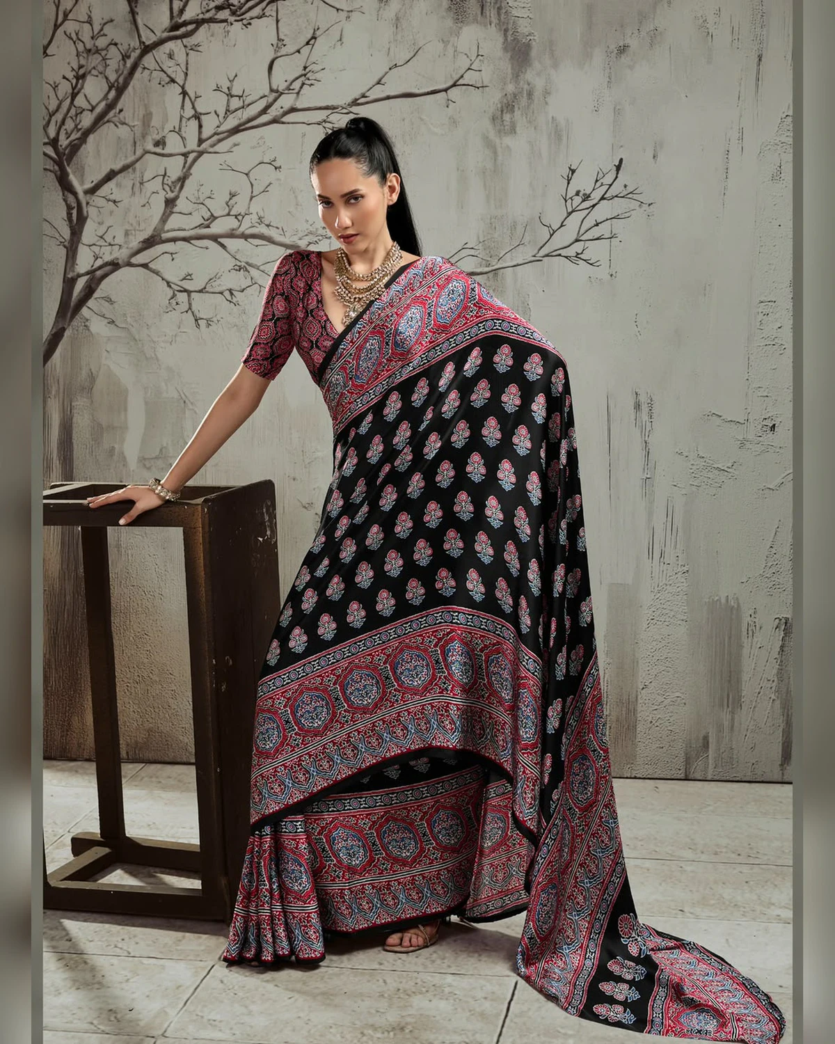Woman wearing a black and red Ajrakh buti modal satin saree with a wide red border, against a grey wall with bare branches