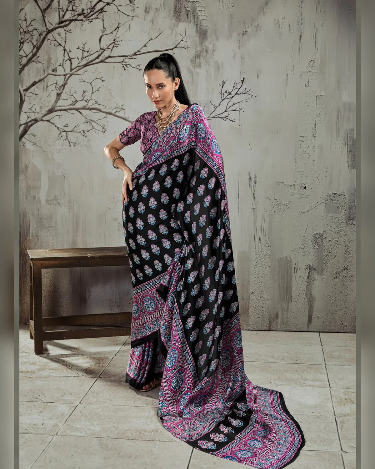Woman wearing a black and purple Ajrakh buti modal satin saree with a wide purple border, against a grey wall with bare branches