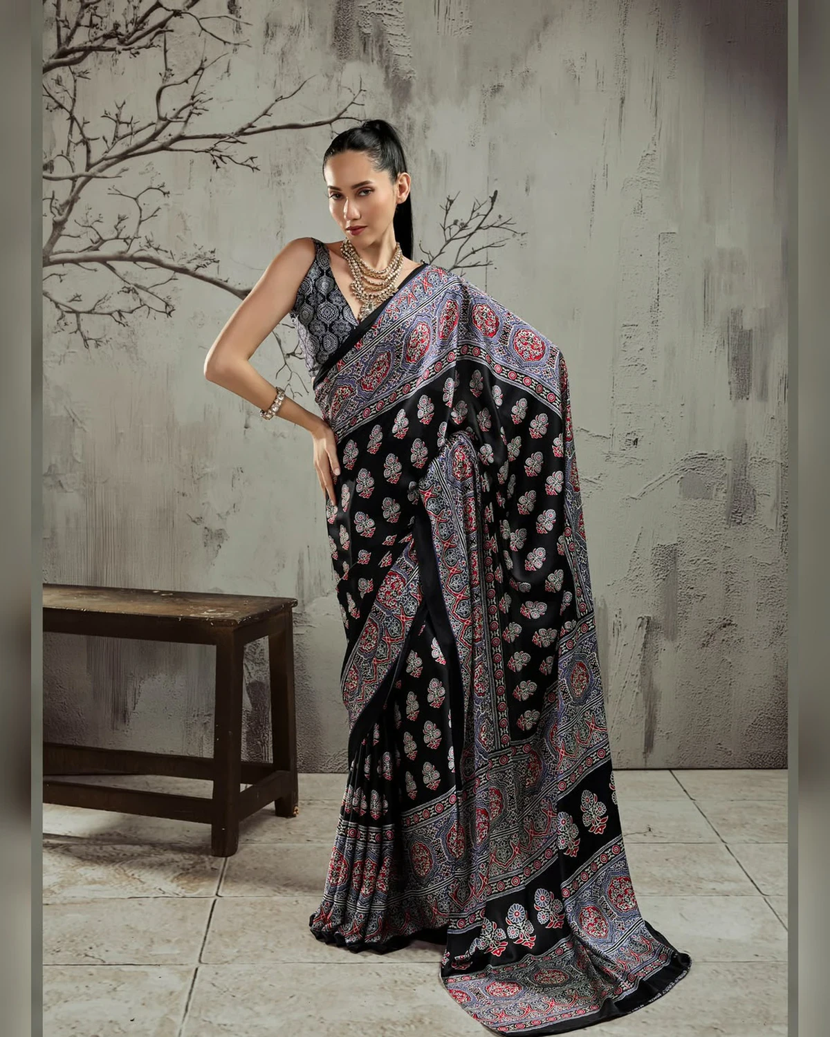 Woman wearing a black and grey Ajrakh buti modal satin saree with a wide grey border, against a grey wall with bare branches