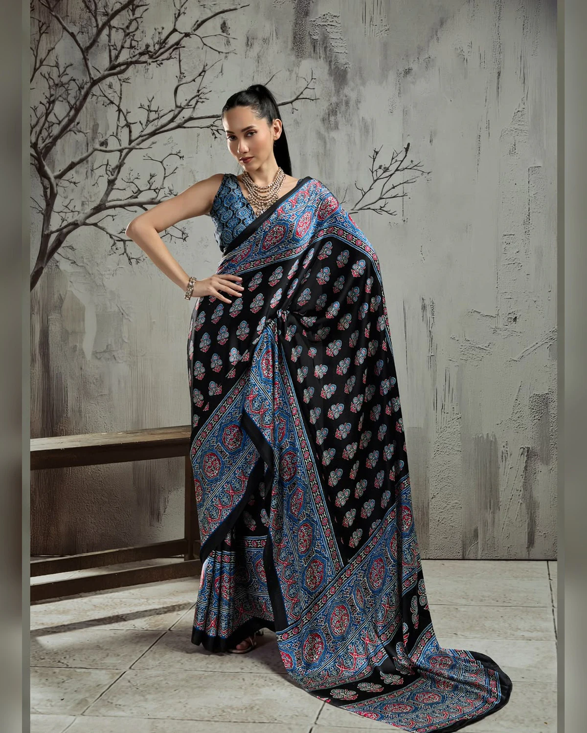 Woman wearing a black and blue Ajrakh buti modal satin saree with a wide blue border, against a grey wall with bare branches