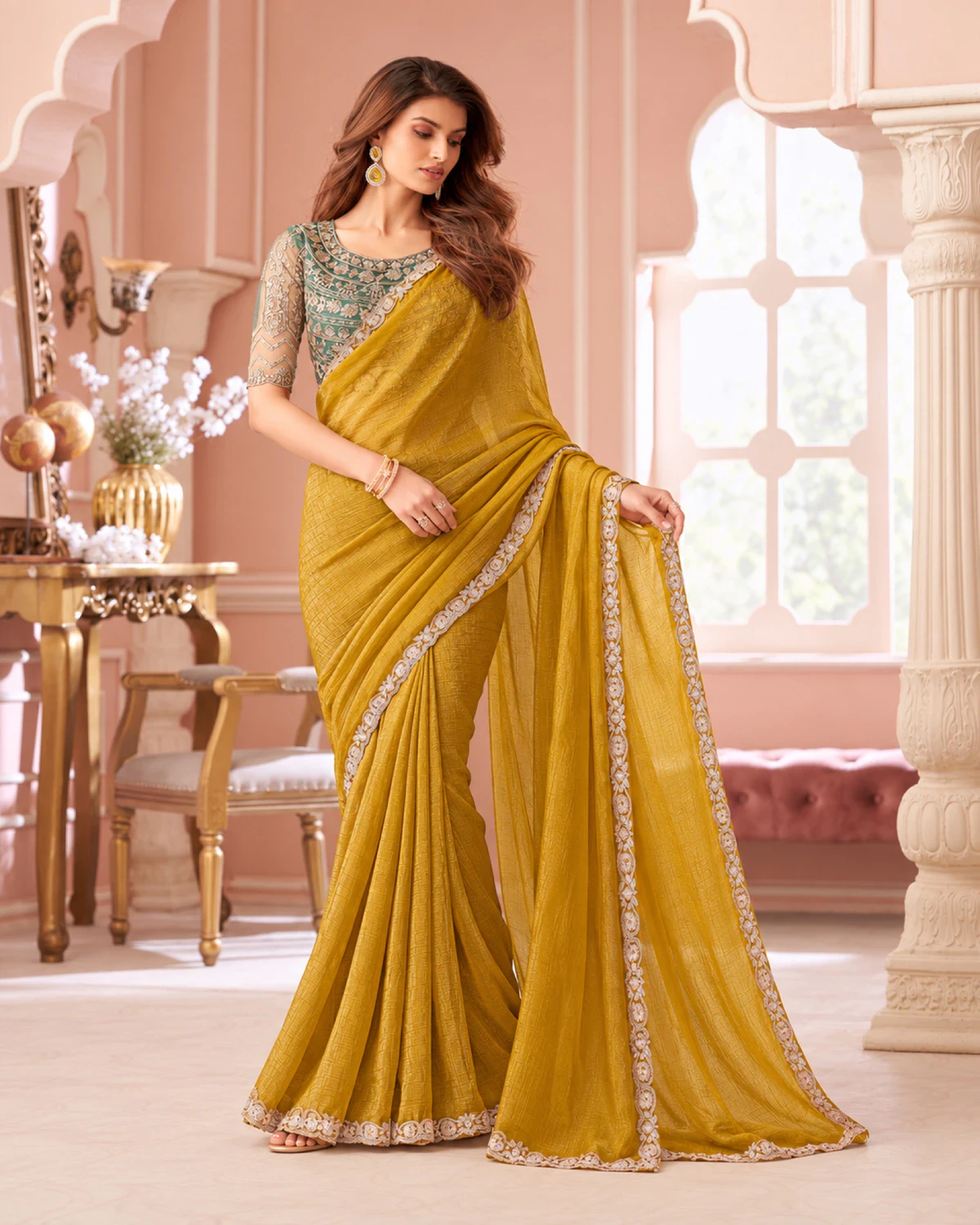 Woman holding out the pallu of the mustard yellow georgette saree in a pink room with arches