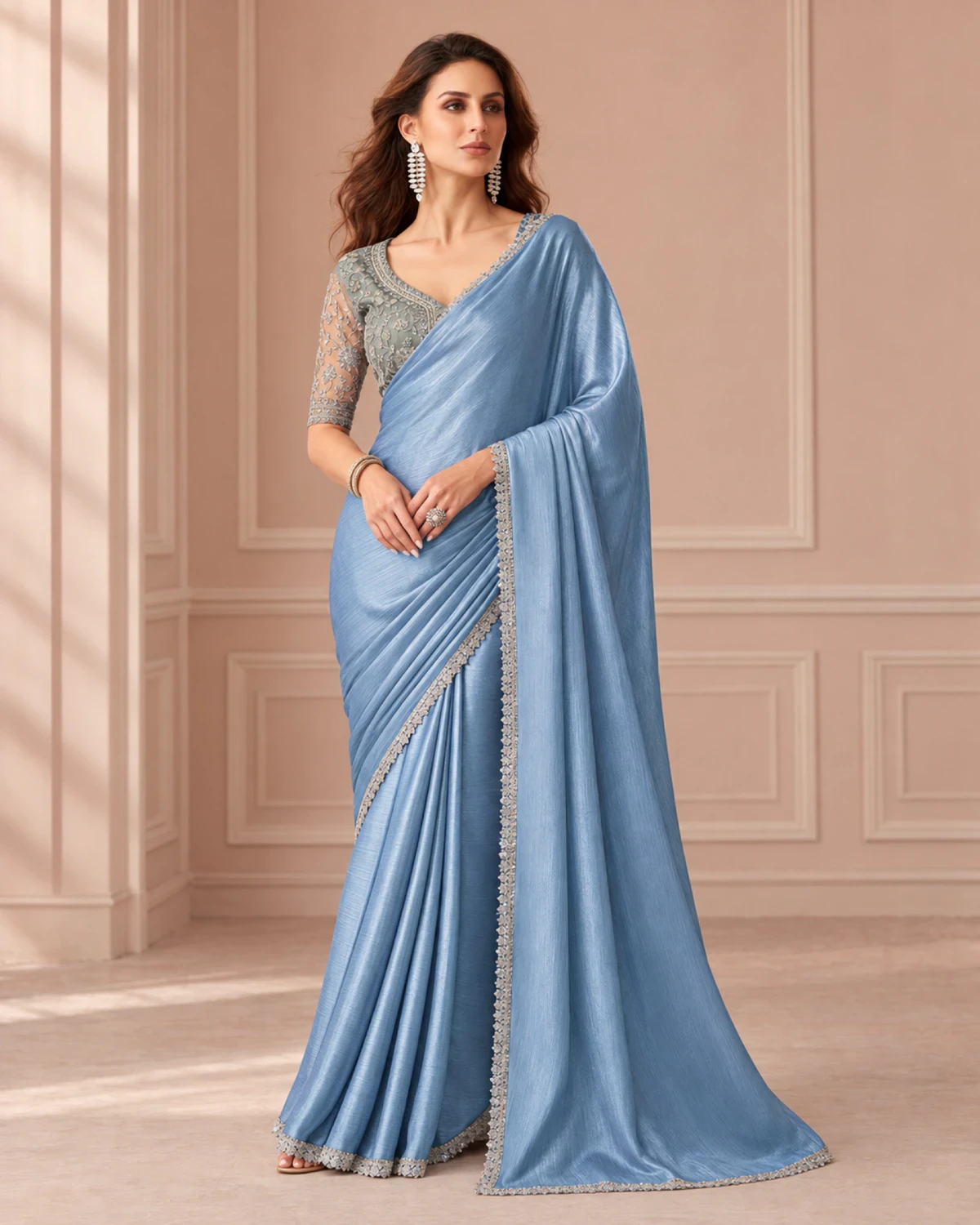 Woman wearing a powder blue shimmer chiffon saree with a scalloped silver border, standing in a softly lit peach room