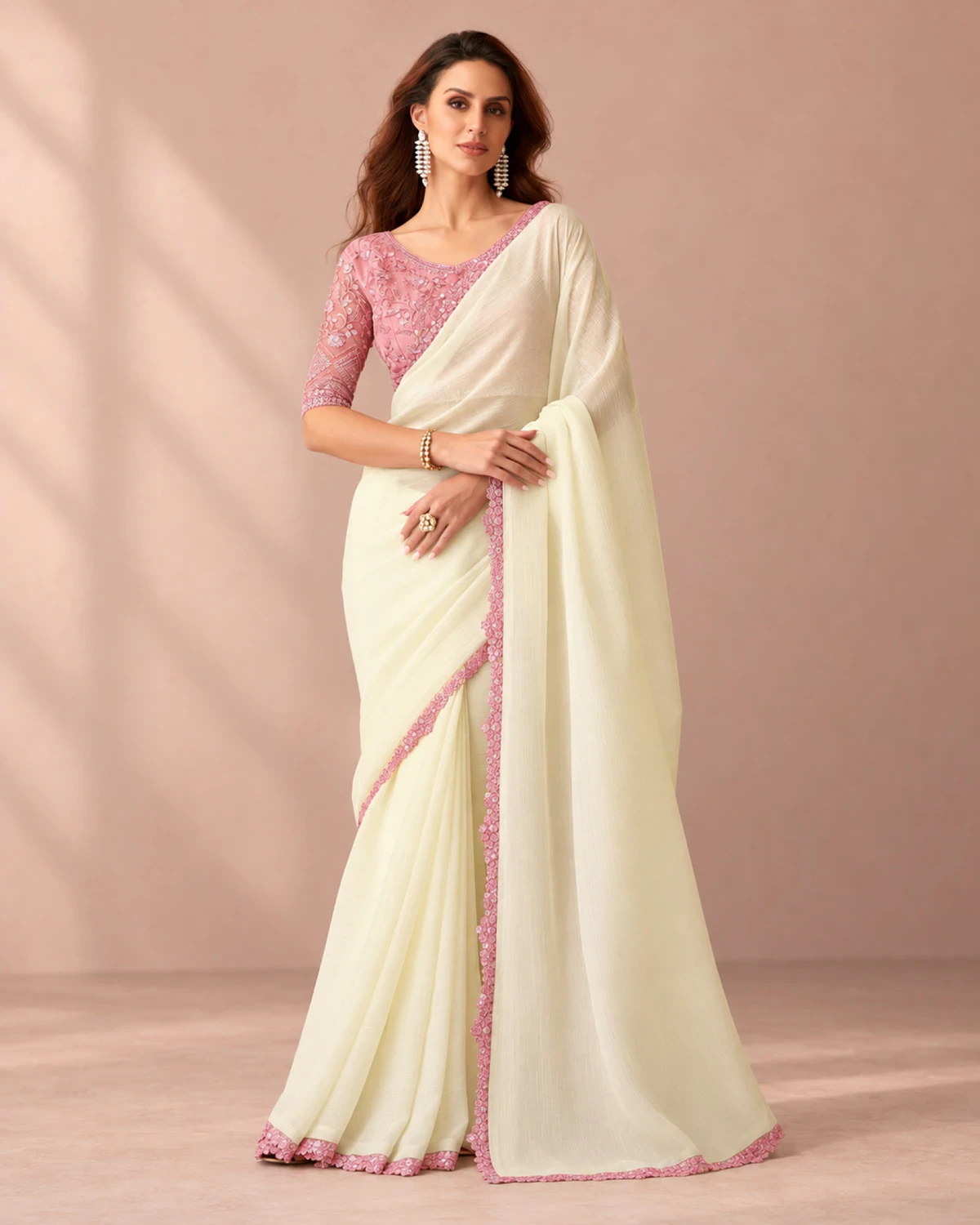 Woman wearing an ivory textured georgette saree with a scalloped pink border, standing in a softly lit peach room