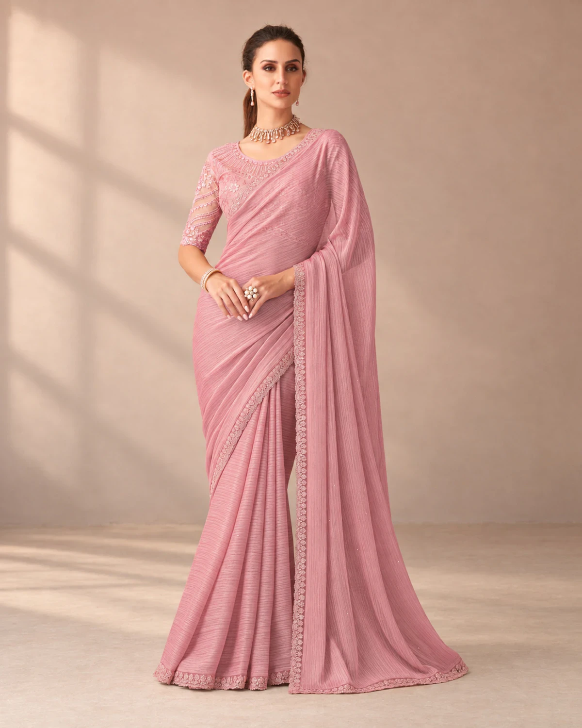 Woman wearing a blush pink textured georgette saree with a scalloped matching border, standing in a softly lit peach room