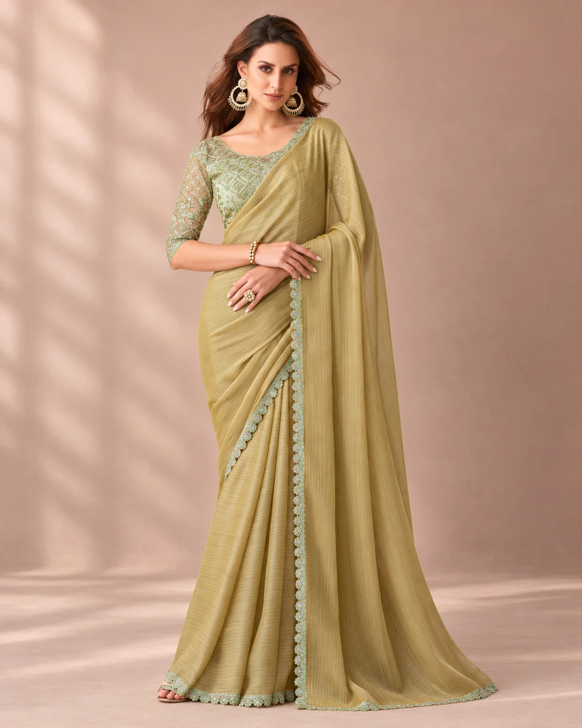 Woman wearing an olive beige textured georgette saree with a scalloped mint green border, standing in a softly lit peach room