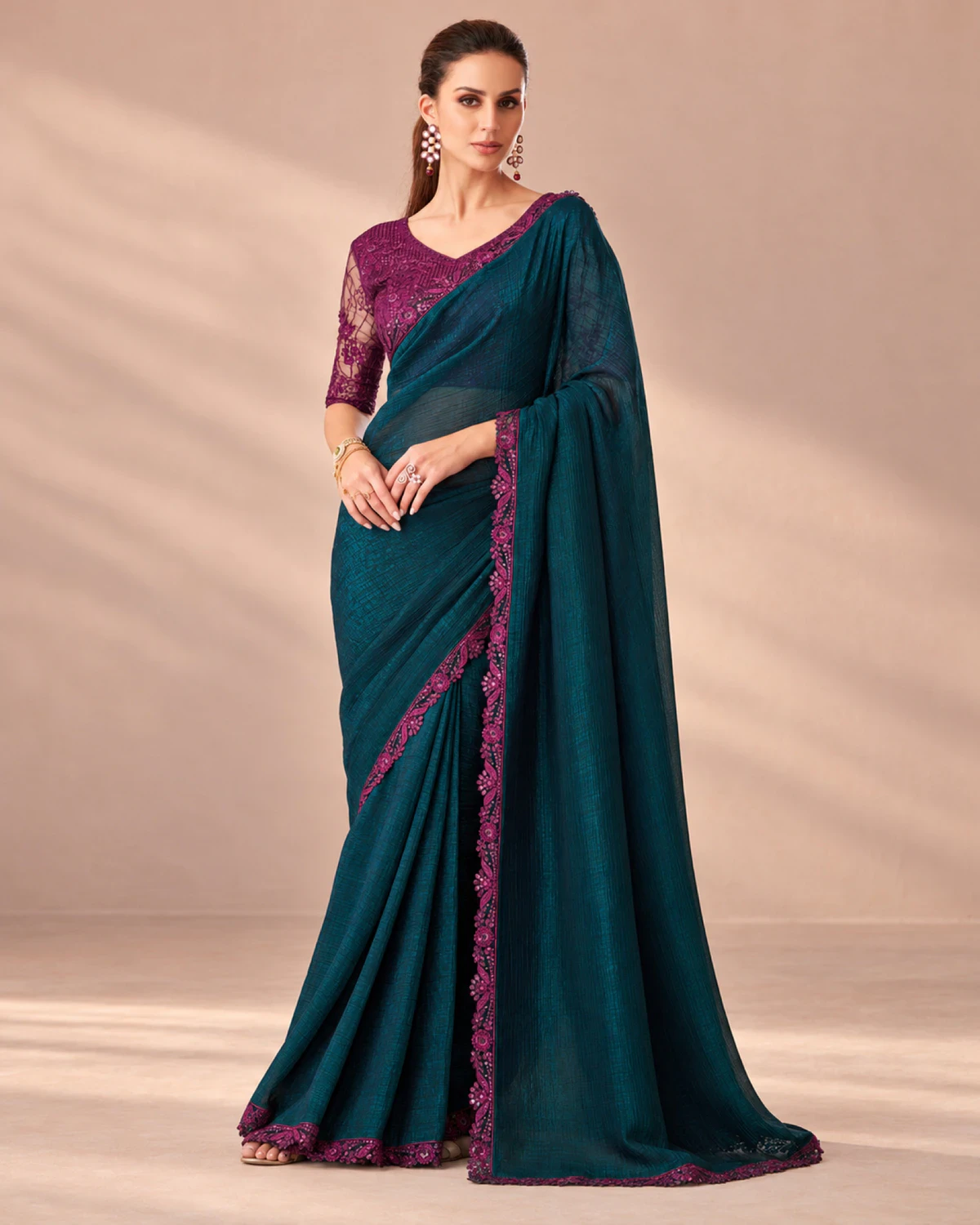 Woman wearing a deep teal peacock rainbow saree with a scalloped magenta border, standing in a softly lit peach room