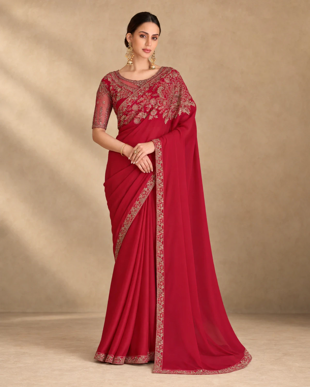 Woman wearing a red sparkle georgette saree with a gold embroidered border, standing against a warm beige backdrop