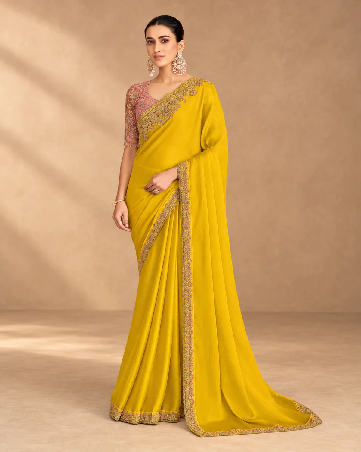 Woman wearing a yellow satin chiffon saree with a gold embroidered border, standing against a warm beige backdrop