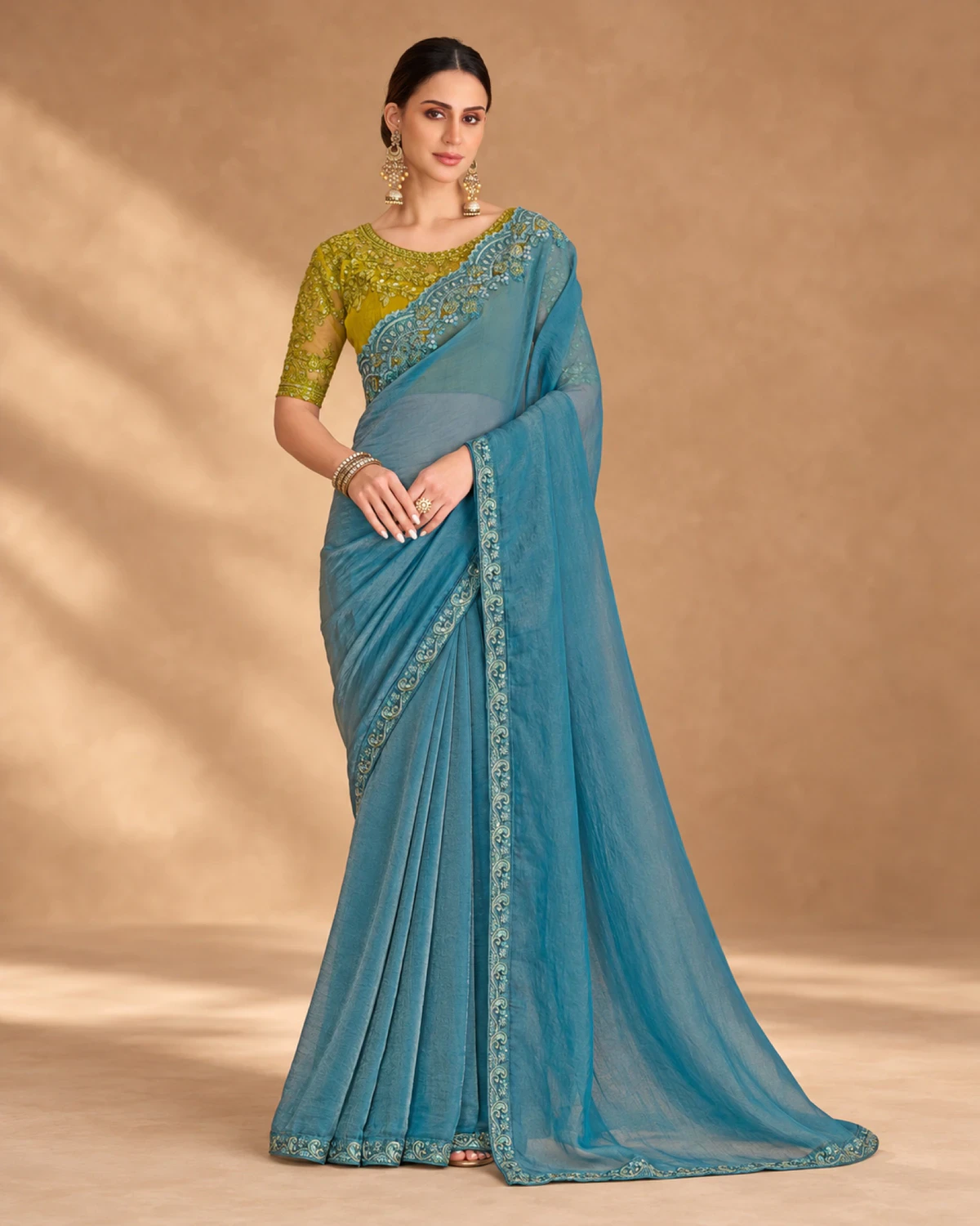 Woman wearing a powder blue tissue georgette saree with an embroidered border, standing against a warm beige backdrop