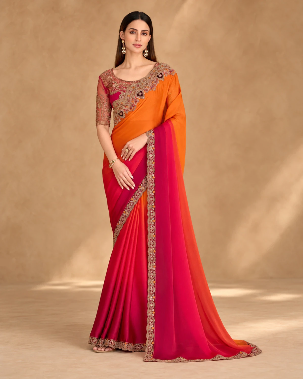 Woman wearing an orange and red shaded satin chiffon saree with a gold embroidered border, standing against a warm beige backdrop