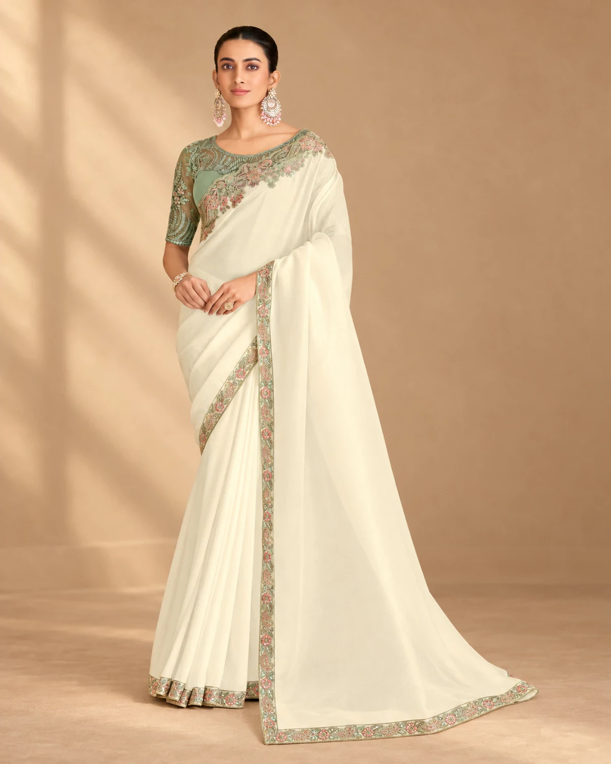 Woman wearing an ivory satin saree with a sage green embroidered border, standing against a warm beige backdrop