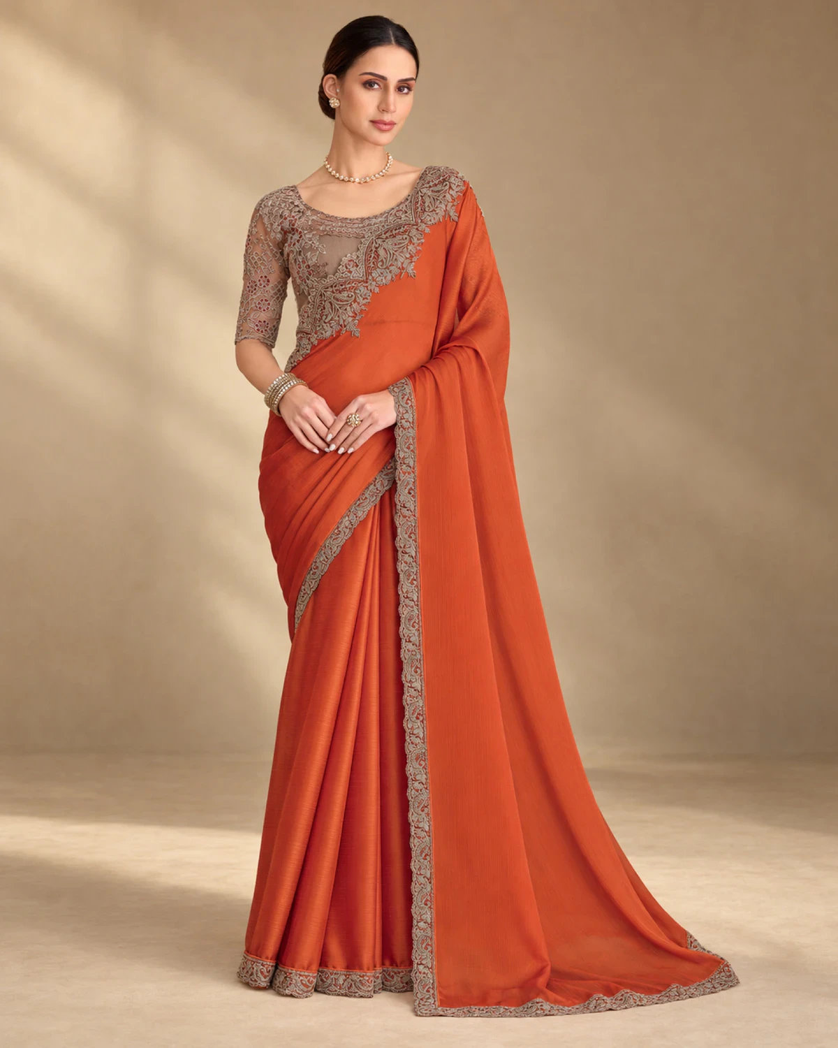 Woman wearing a rust satin chiffon saree with a copper-toned embroidered border, standing against a warm beige backdrop
