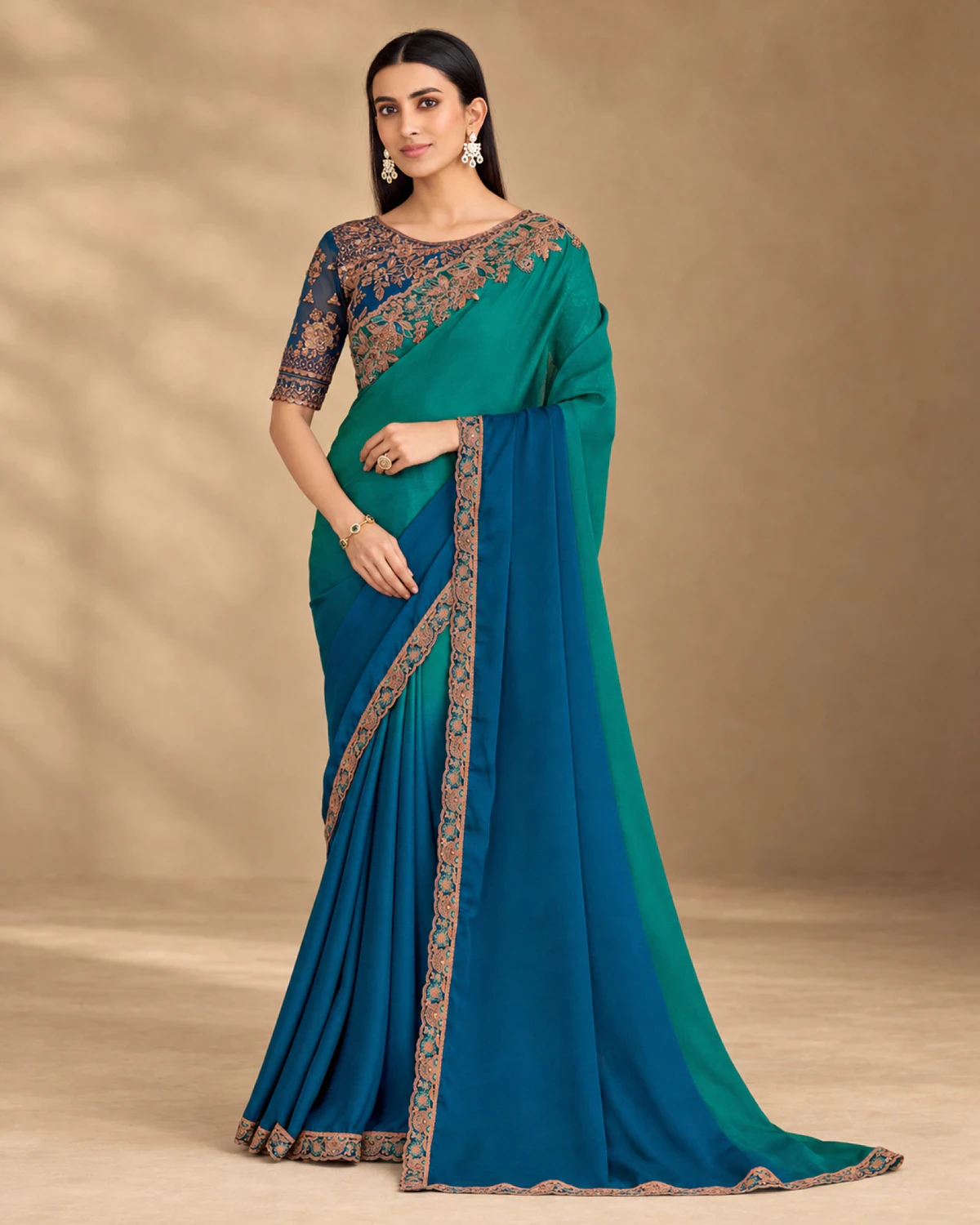 Woman wearing a teal and blue shaded chiffon saree with a copper embroidered border, standing against a warm beige backdrop