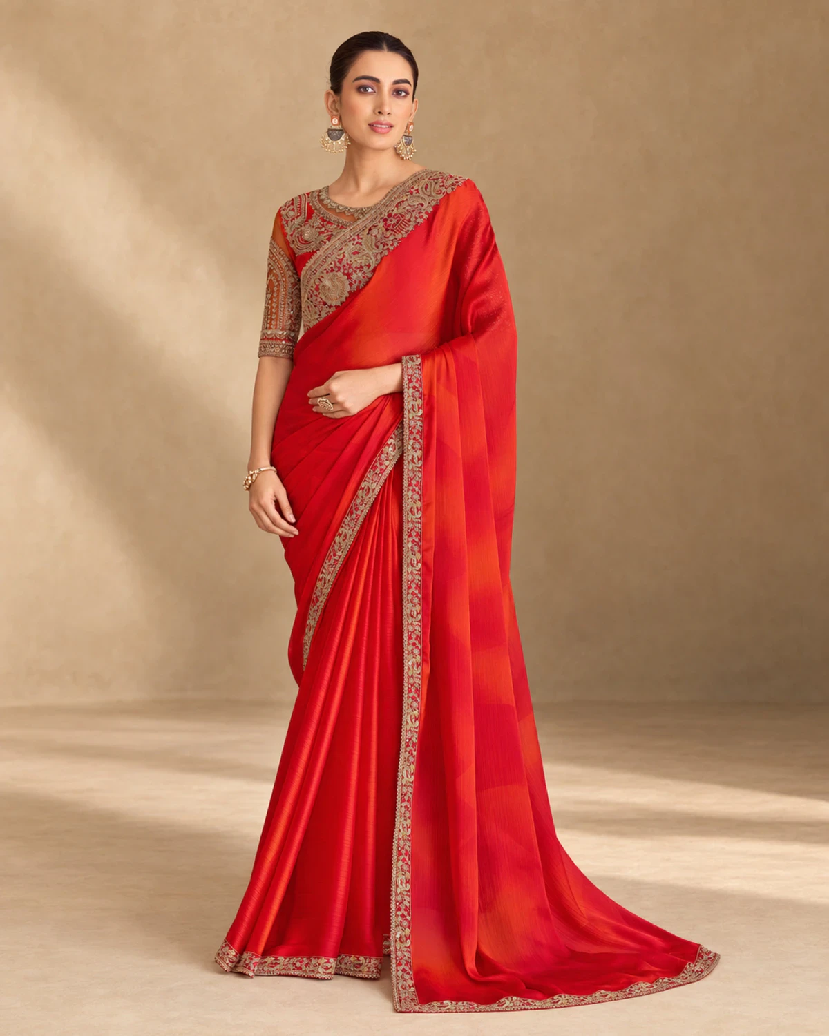 Woman wearing a red shaded sparkle chiffon saree with a gold embroidered border, standing against a warm beige backdrop
