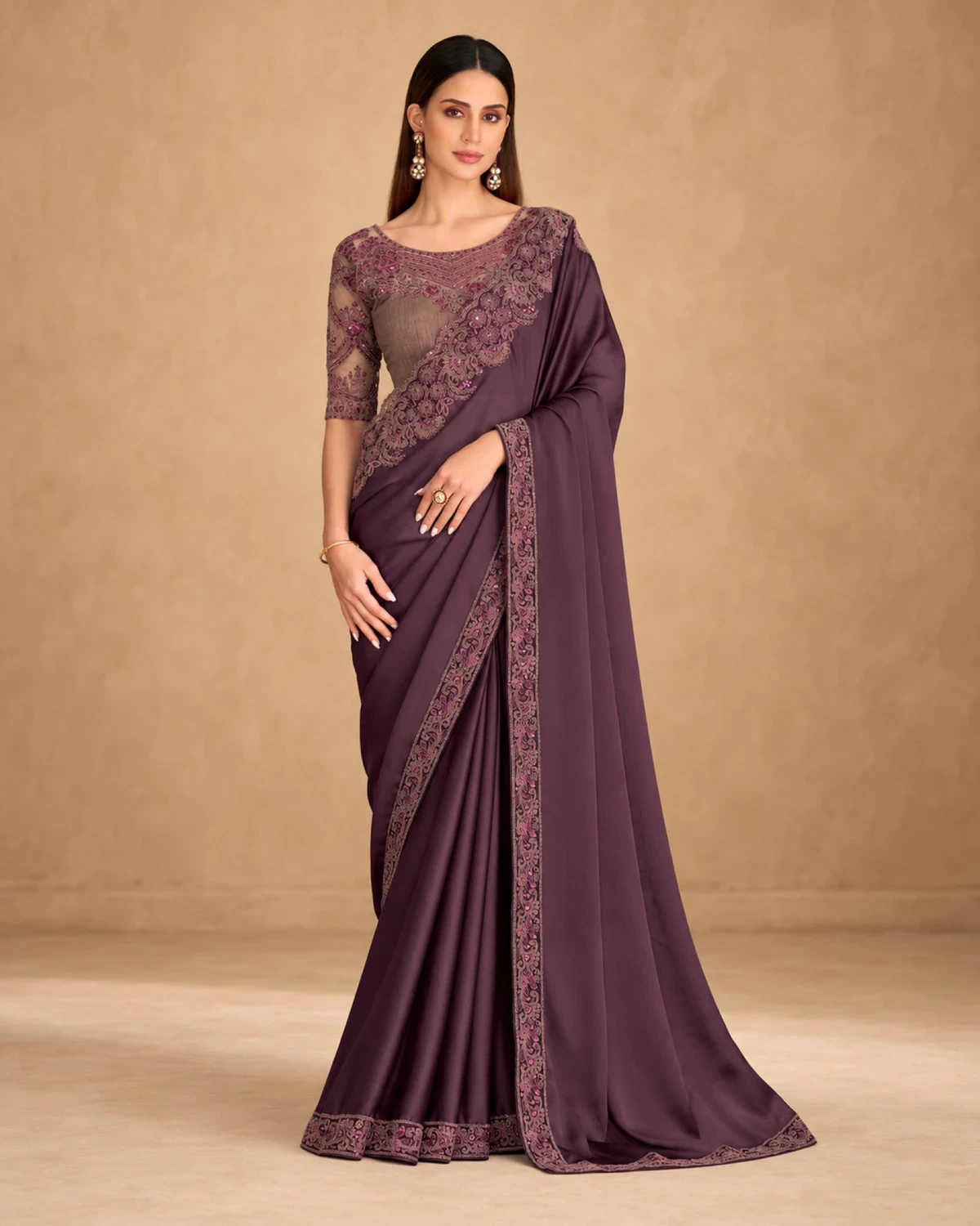 Woman wearing a plum satin chiffon saree with a mauve embroidered border, standing against a warm beige backdrop