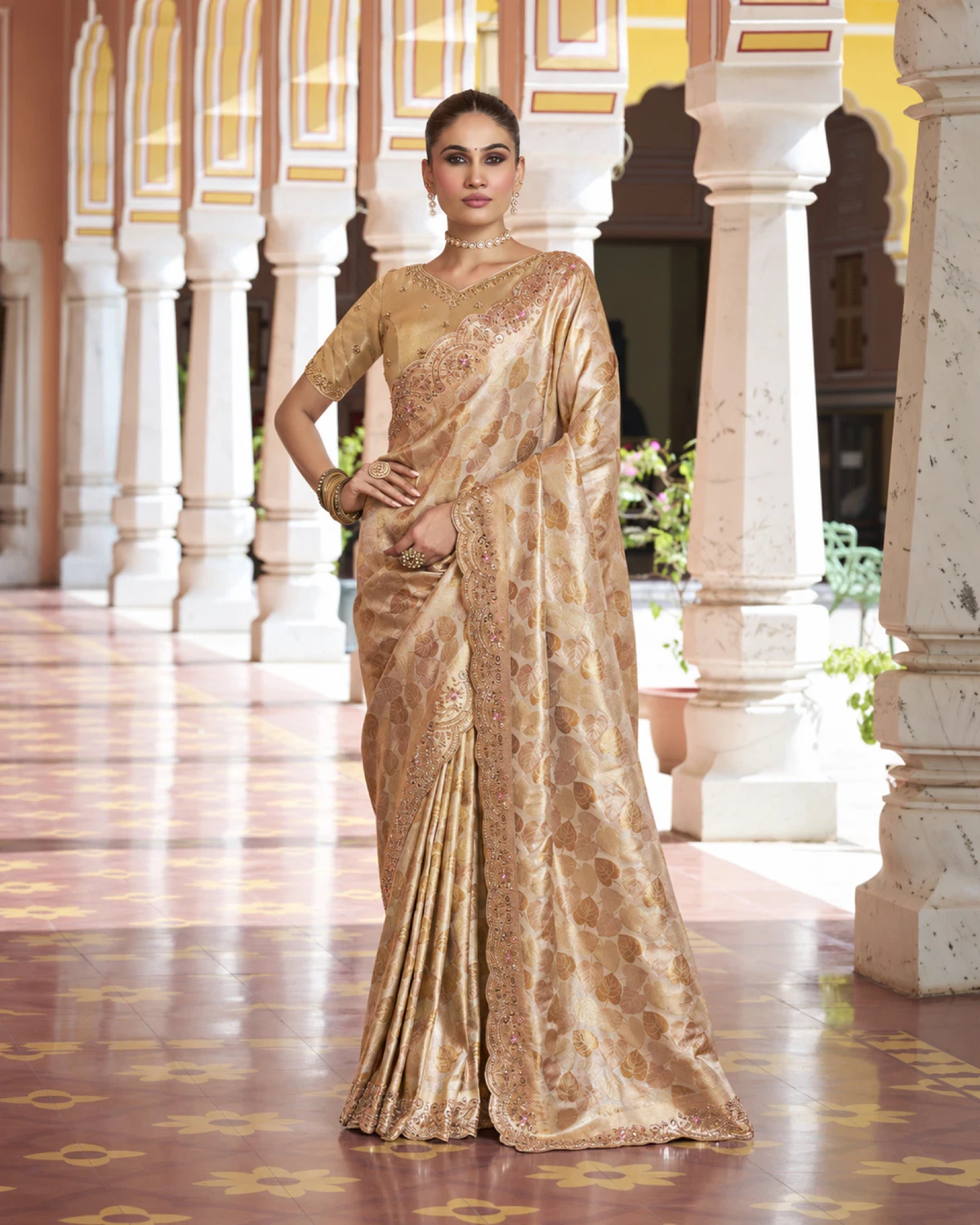 Woman wearing a champagne gold Kanjivaram-style soft silk saree with a scalloped beaded border, standing in a pillared courtyard
