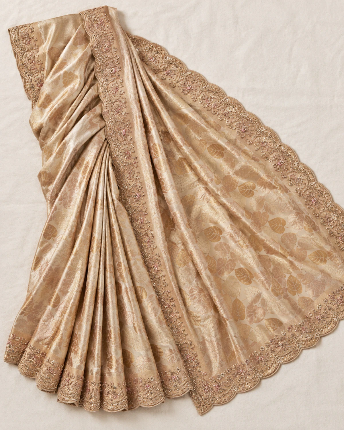 The champagne gold soft silk saree laid flat on a cream cloth, showing its woven gold leaves and scalloped border