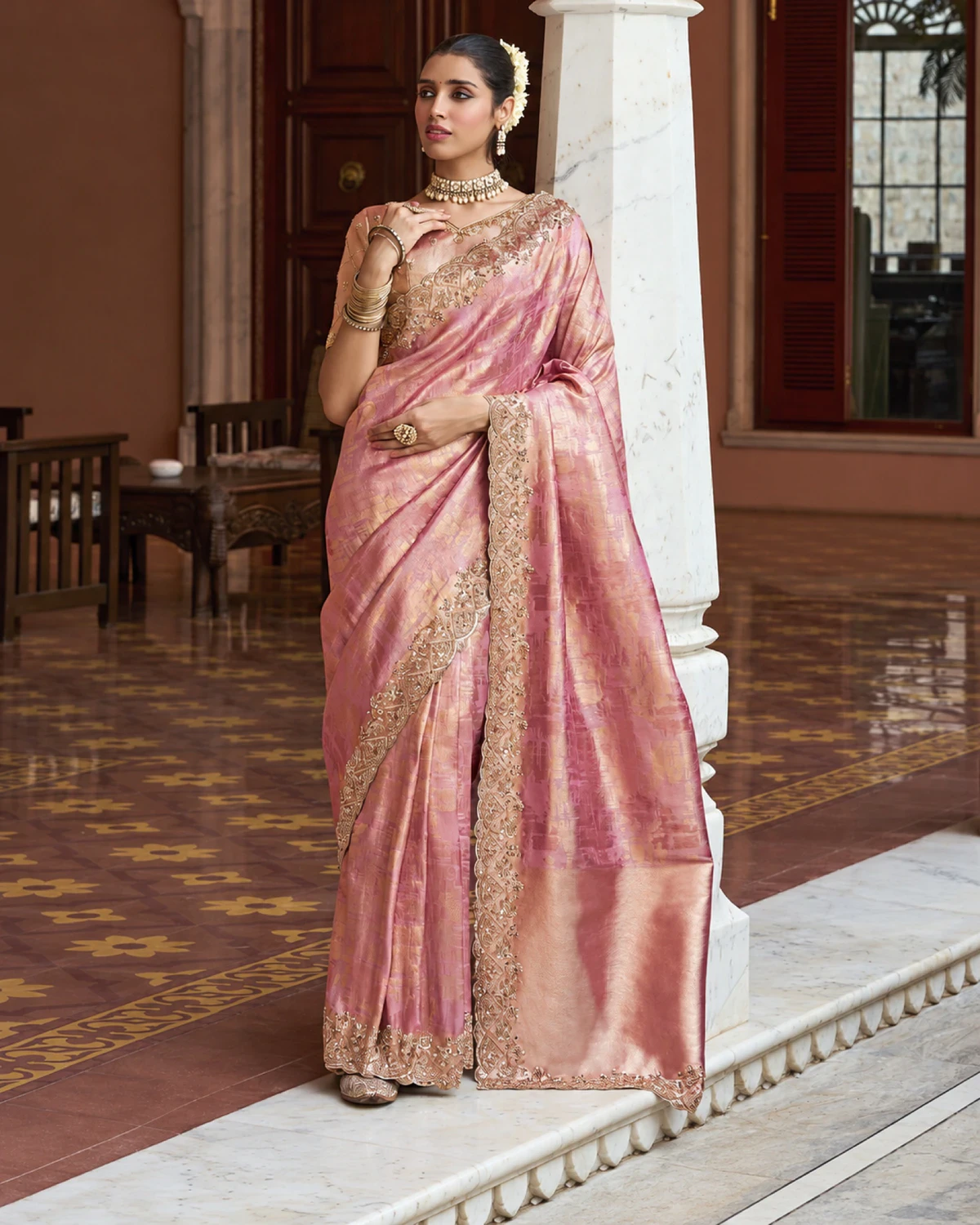 Woman wearing a rose pink Kanjivaram-style soft silk saree with a beaded scalloped border, standing by a white pillar