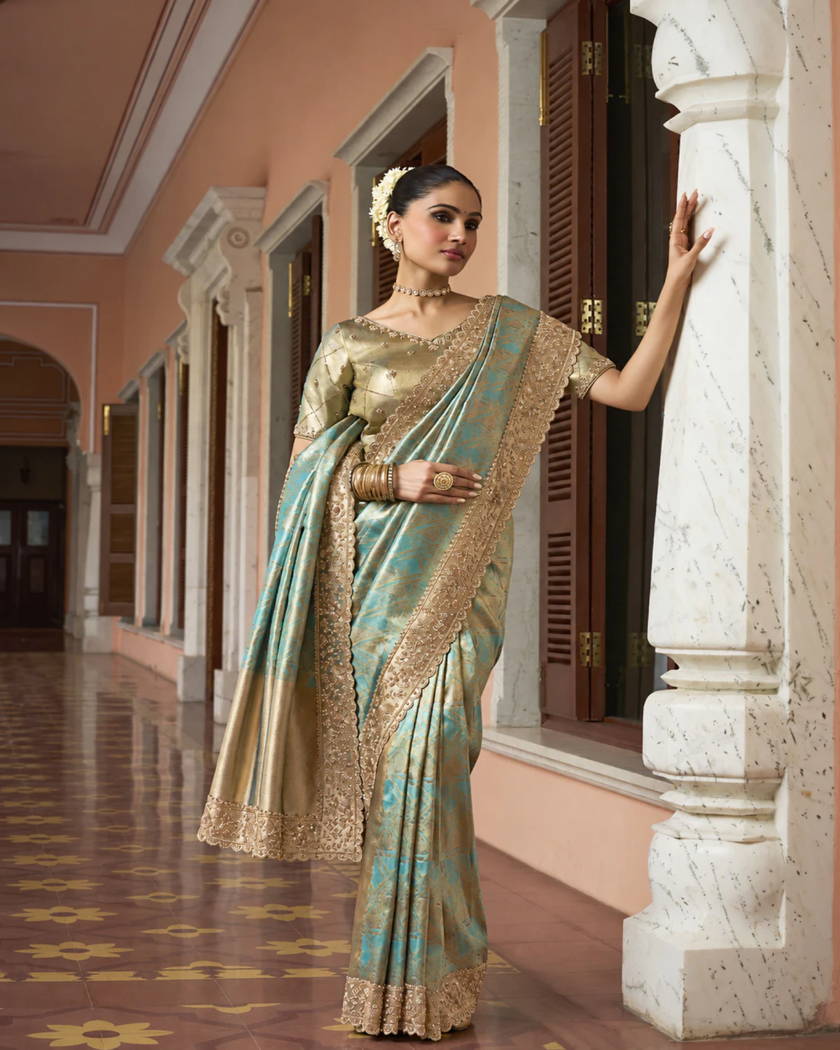 Woman wearing an aqua Kanjivaram-style soft silk saree with a wide beaded border, standing by a marble pillar