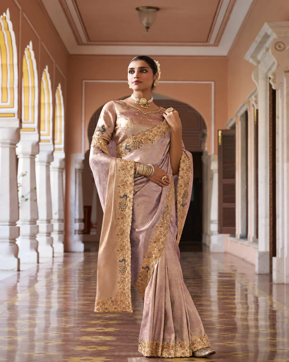 Woman wearing a dusty lilac Kanjivaram-style soft silk saree with a gold peacock border, in a long arched corridor