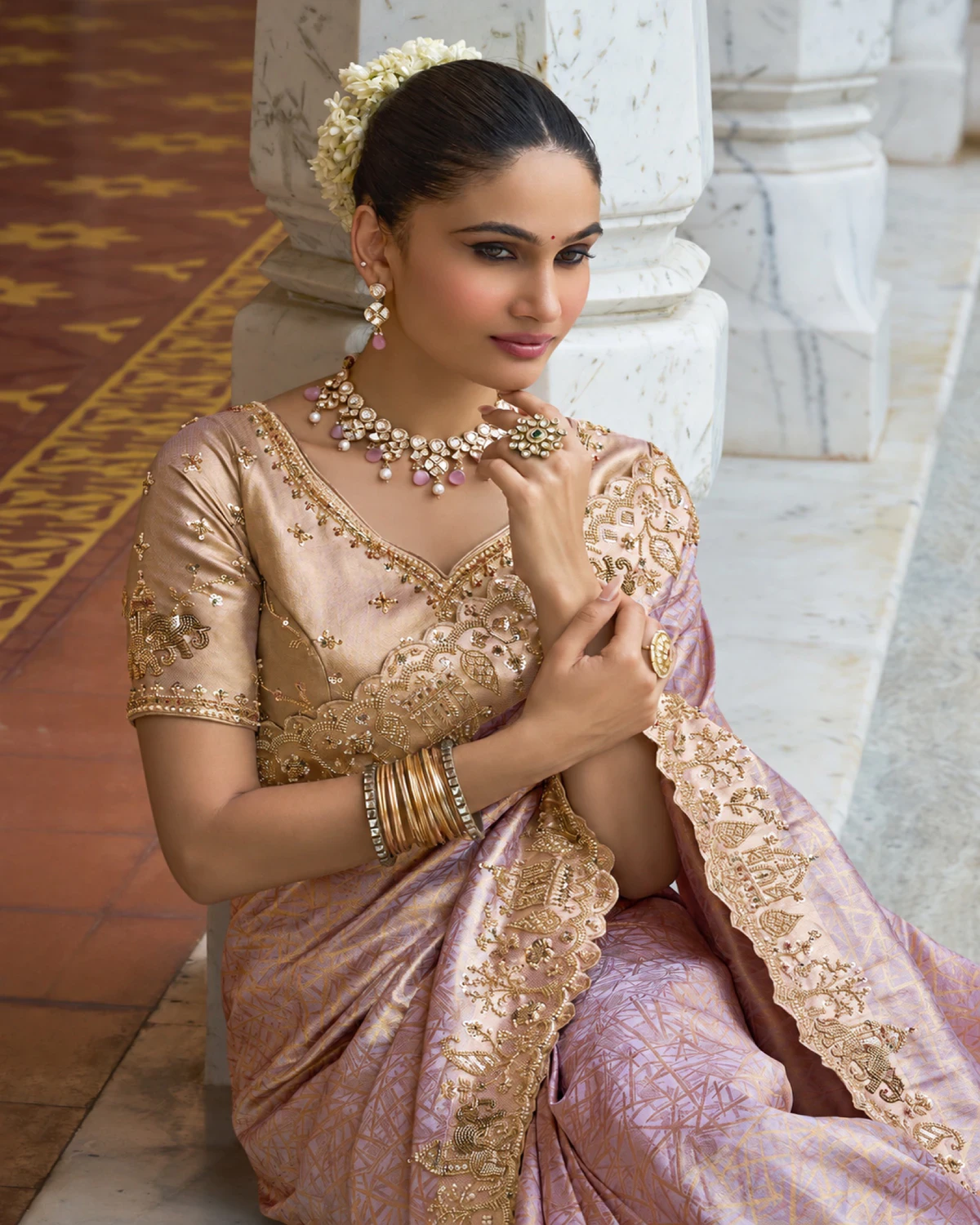 Woman seated by a pillar in the blush lilac soft silk saree, showing its beaded border and blouse